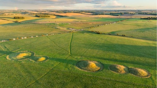 A wide aerial shot of the Stonehenge Landscape with The Cursus Barrows in the foreground and the monument visible further away in the distance.